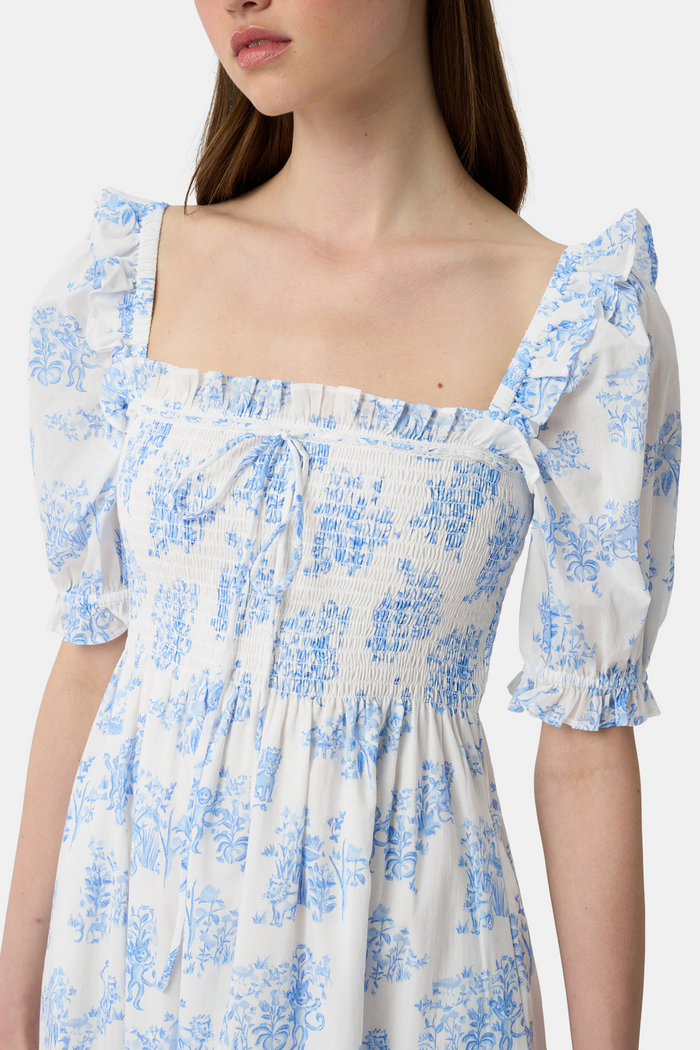 A woman wears the Roller Rabbit Sherwood Forest Monkey Scarlett Nap Dress—long, white with a blue monkey toile print, puffed short sleeves, smocked bodice, and square neckline—paired with blue slip-on sandals against a light background.