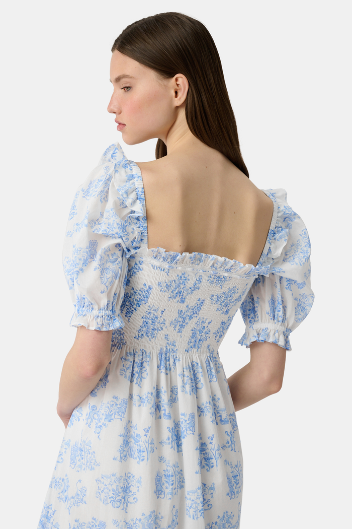 A woman wears the Roller Rabbit Sherwood Forest Monkey Scarlett Nap Dress—long, white with a blue monkey toile print, puffed short sleeves, smocked bodice, and square neckline—paired with blue slip-on sandals against a light background.