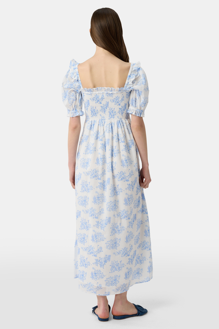 A woman wears the Roller Rabbit Sherwood Forest Monkey Scarlett Nap Dress—long, white with a blue monkey toile print, puffed short sleeves, smocked bodice, and square neckline—paired with blue slip-on sandals against a light background.