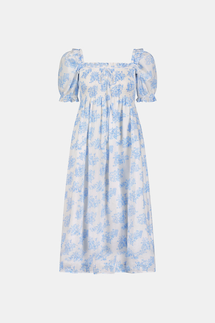 A woman wears the Roller Rabbit Sherwood Forest Monkey Scarlett Nap Dress—long, white with a blue monkey toile print, puffed short sleeves, smocked bodice, and square neckline—paired with blue slip-on sandals against a light background.