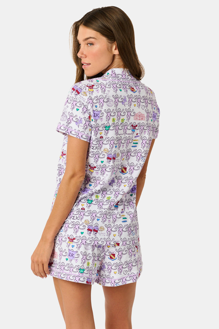 The Sigma Kappa Monkey Polo Pajamas by Roller Rabbit are a short-sleeve pajama set featuring matching shorts with a white background and colorful cartoon character pattern. Designed for comfort, they include a front button placket and an elastic waistband.