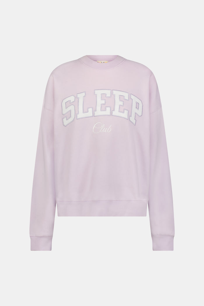 A woman wearing the Roller Rabbit Sleep Club Andie Sweatshirt in pastel lavender, paired with patterned white shorts, stands and smiles against a plain white background.