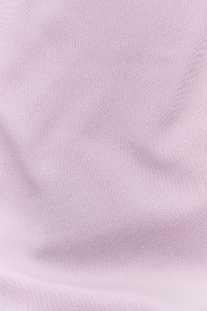 A woman wearing the Roller Rabbit Sleep Club Andie Sweatshirt in pastel lavender, paired with patterned white shorts, stands and smiles against a plain white background.