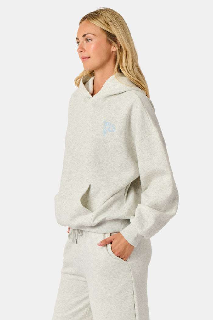 A person is wearing the Roller Rabbit Sleep Club Mimi Sweatpants in light gray, featuring wide legs and crafted from velvety soft Dream Fleece with “Sleep Club” printed in light blue on the left thigh, paired with white sneakers and a cropped white top. The background is plain white.
