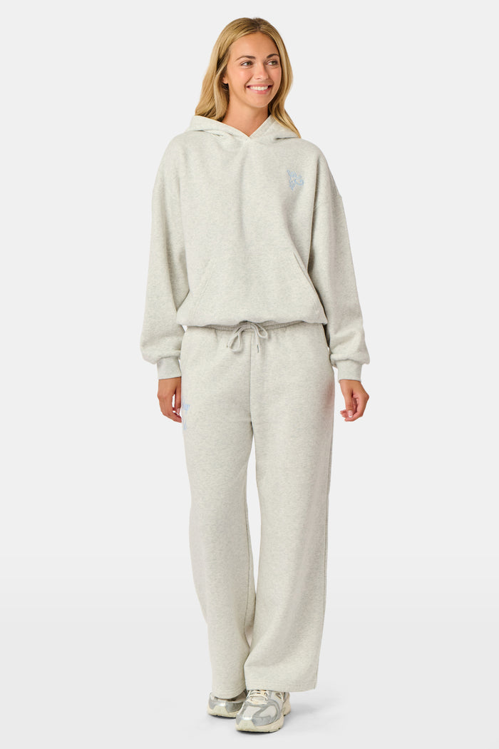 A person is wearing the Roller Rabbit Sleep Club Mimi Sweatpants in light gray, featuring wide legs and crafted from velvety soft Dream Fleece with “Sleep Club” printed in light blue on the left thigh, paired with white sneakers and a cropped white top. The background is plain white.