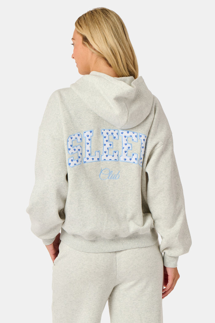 A person is wearing the Roller Rabbit Sleep Club Mimi Sweatpants in light gray, featuring wide legs and crafted from velvety soft Dream Fleece with “Sleep Club” printed in light blue on the left thigh, paired with white sneakers and a cropped white top. The background is plain white.