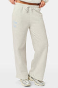 A person is wearing the Roller Rabbit Sleep Club Mimi Sweatpants in light gray, featuring wide legs and crafted from velvety soft Dream Fleece with “Sleep Club” printed in light blue on the left thigh, paired with white sneakers and a cropped white top. The background is plain white.