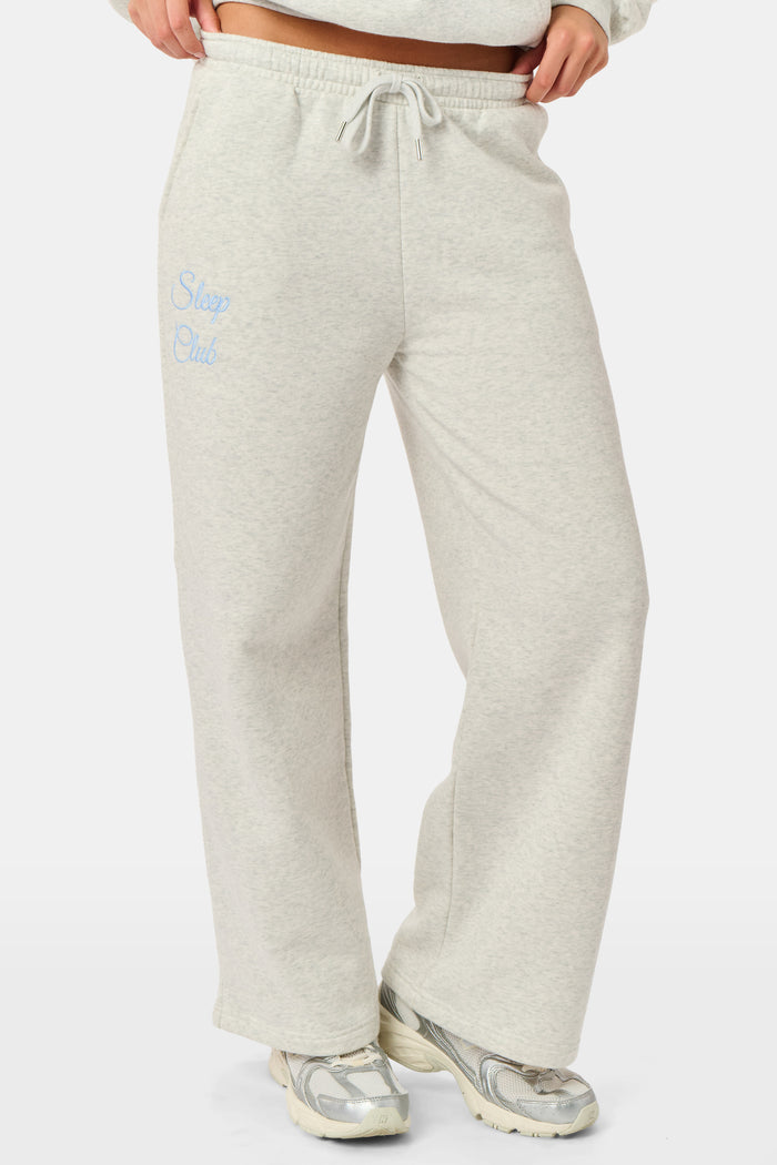 A person is wearing the Roller Rabbit Sleep Club Mimi Sweatpants in light gray, featuring wide legs and crafted from velvety soft Dream Fleece with “Sleep Club” printed in light blue on the left thigh, paired with white sneakers and a cropped white top. The background is plain white.