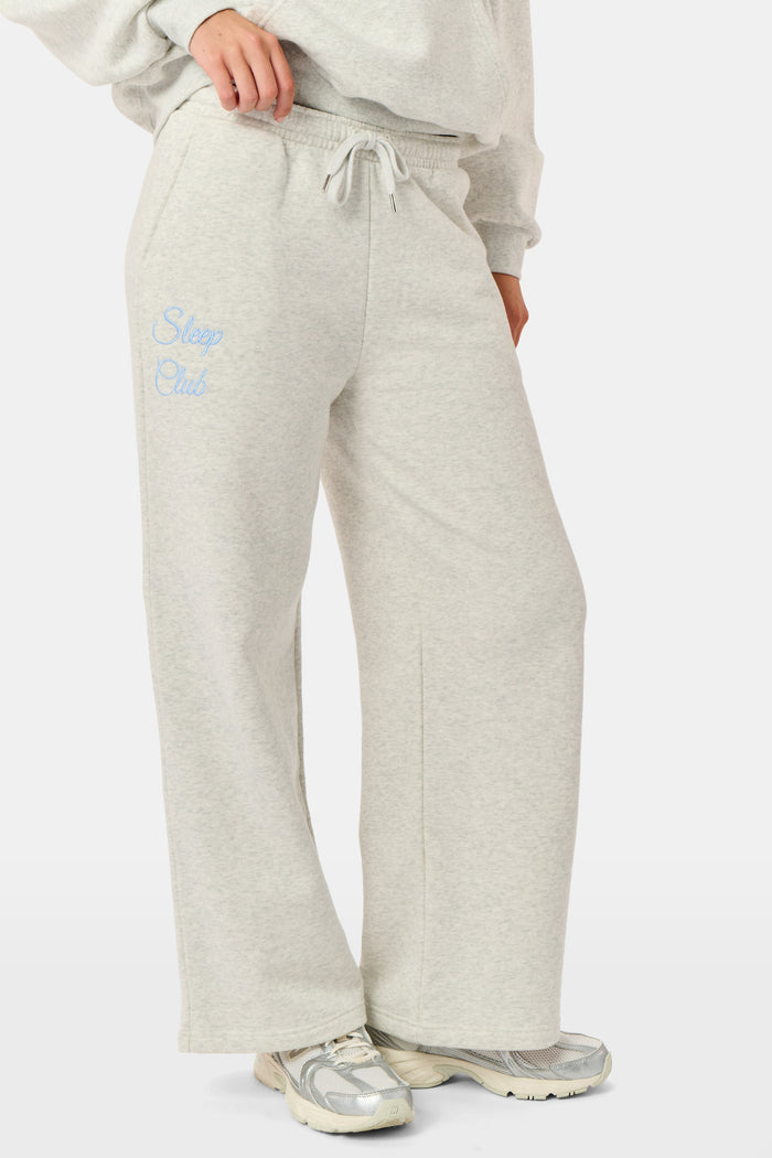 A person is wearing the Roller Rabbit Sleep Club Mimi Sweatpants in light gray, featuring wide legs and crafted from velvety soft Dream Fleece with “Sleep Club” printed in light blue on the left thigh, paired with white sneakers and a cropped white top. The background is plain white.