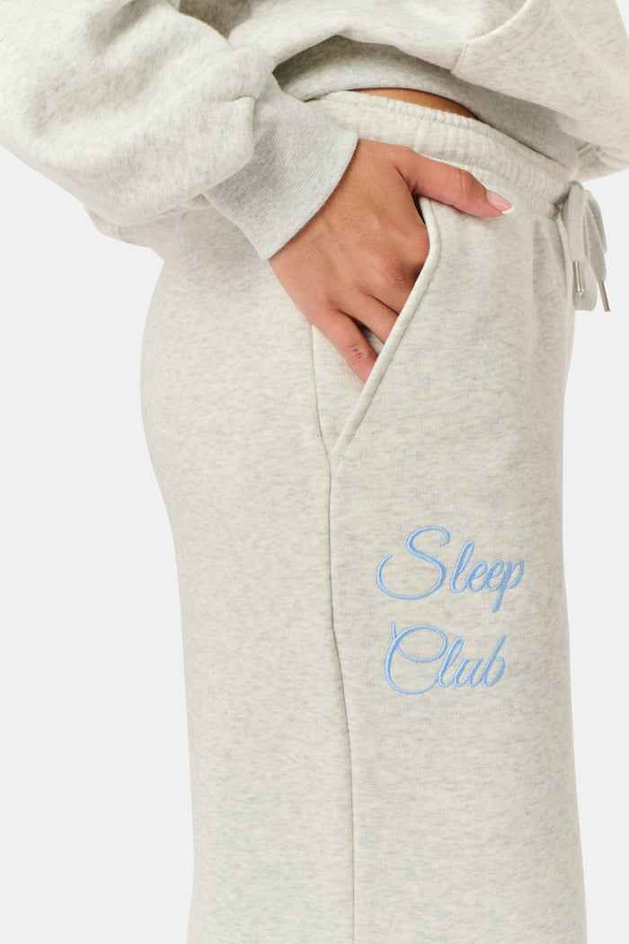 A person is wearing the Roller Rabbit Sleep Club Mimi Sweatpants in light gray, featuring wide legs and crafted from velvety soft Dream Fleece with “Sleep Club” printed in light blue on the left thigh, paired with white sneakers and a cropped white top. The background is plain white.