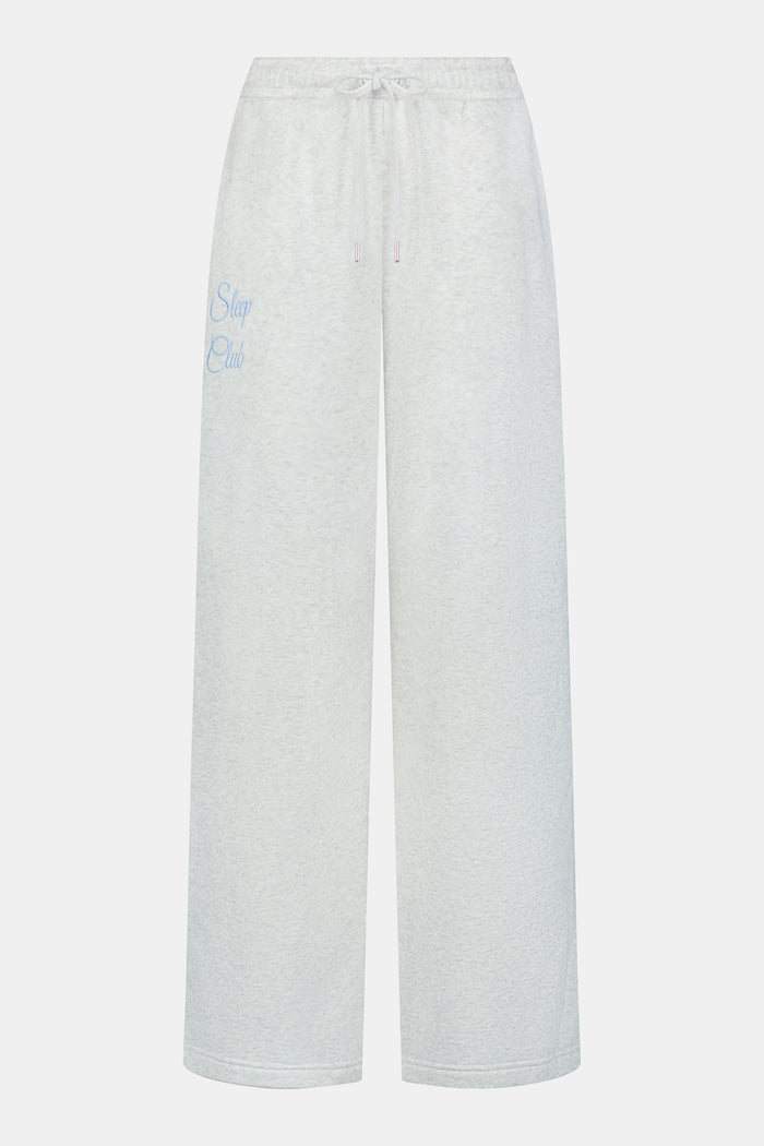 A person is wearing the Roller Rabbit Sleep Club Mimi Sweatpants in light gray, featuring wide legs and crafted from velvety soft Dream Fleece with “Sleep Club” printed in light blue on the left thigh, paired with white sneakers and a cropped white top. The background is plain white.