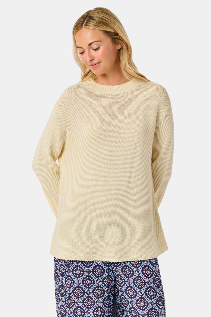 A woman wearing the Roller Rabbit Soleil Sweater, a cream cashmere blend pullover with wide sleeves, paired with patterned blue and white pants, stands against a plain white background.