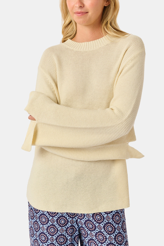 A woman wearing the Roller Rabbit Soleil Sweater, a cream cashmere blend pullover with wide sleeves, paired with patterned blue and white pants, stands against a plain white background.