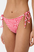 A person wears the Roller Rabbit Starry Dot Isa Bikini Bottom in red with white polka dots and adjustable hip ties, shown from mid-torso to upper thigh.