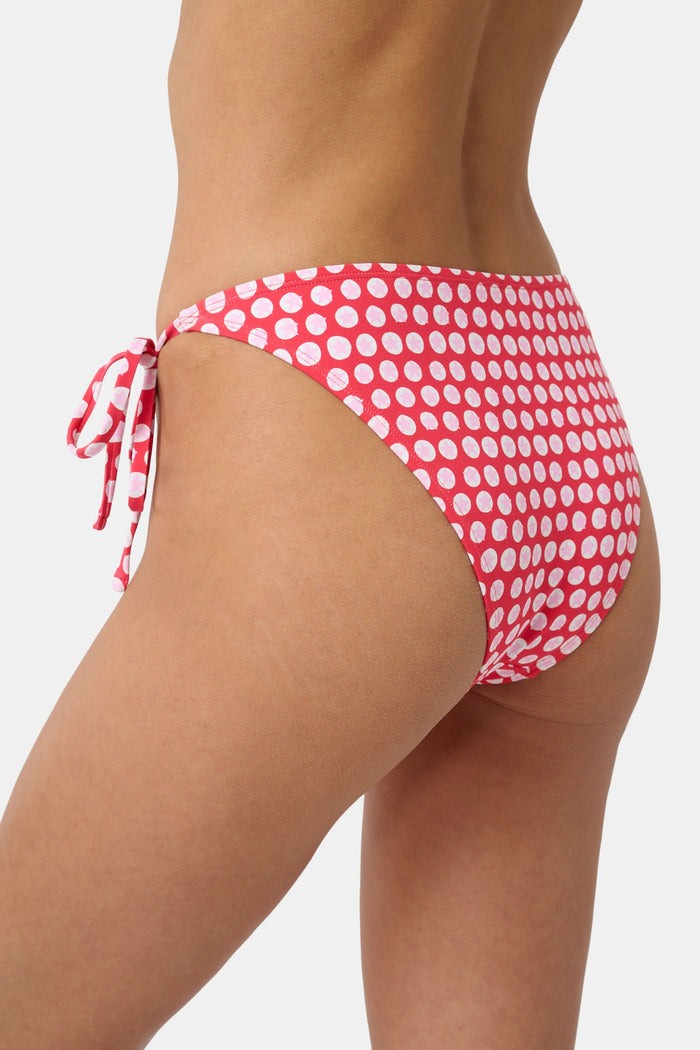 A person wears the Roller Rabbit Starry Dot Isa Bikini Bottom in red with white polka dots and adjustable hip ties, shown from mid-torso to upper thigh.
