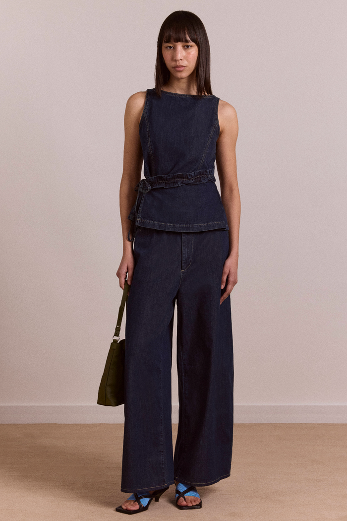 Woman wearing a sleeveless denim top and wide-leg jeans against a plain background