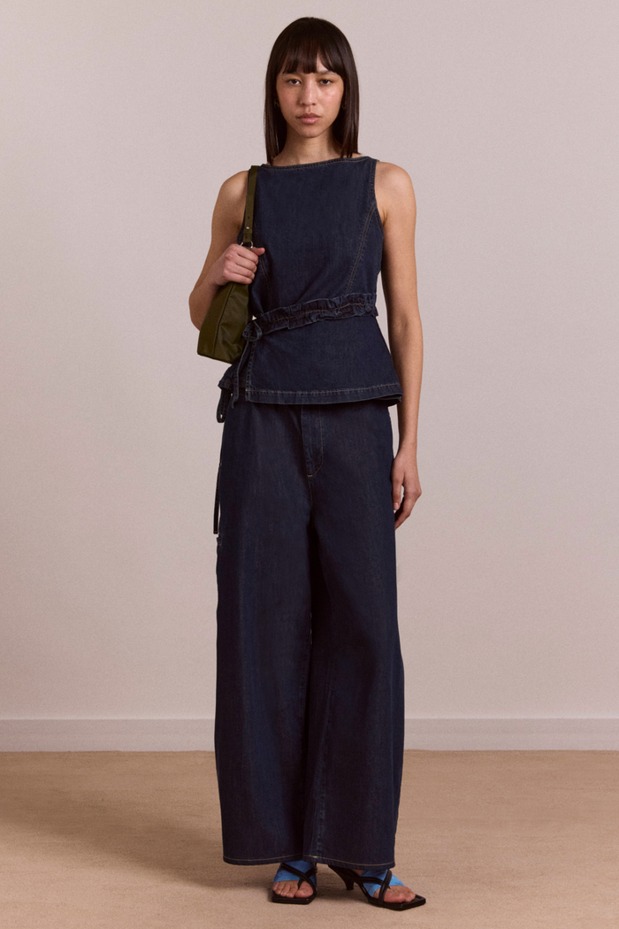 Woman wearing a navy sleeveless top and wide-leg pants holding a green bag against a plain background