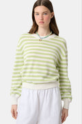 A woman with straight brown hair wears the Roller Rabbit Striped Hadley Sweater in sage green and white stripes, paired with white high-waisted pants and a beaded necklace, standing against a plain background and looking at the camera.