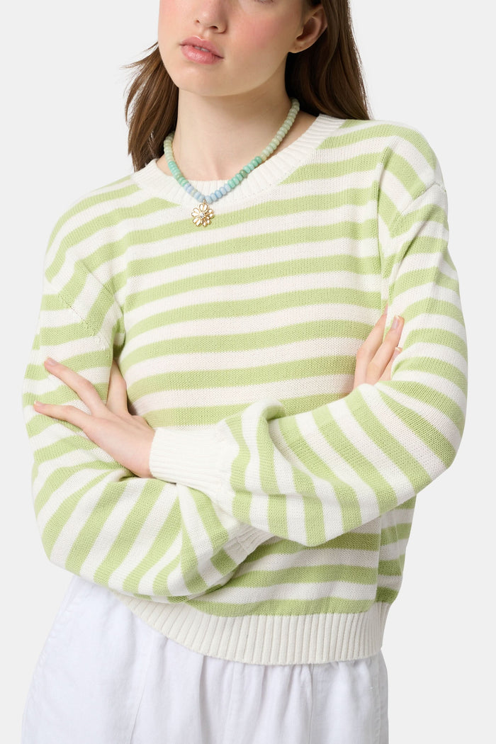 A woman with straight brown hair wears the Roller Rabbit Striped Hadley Sweater in sage green and white stripes, paired with white high-waisted pants and a beaded necklace, standing against a plain background and looking at the camera.
