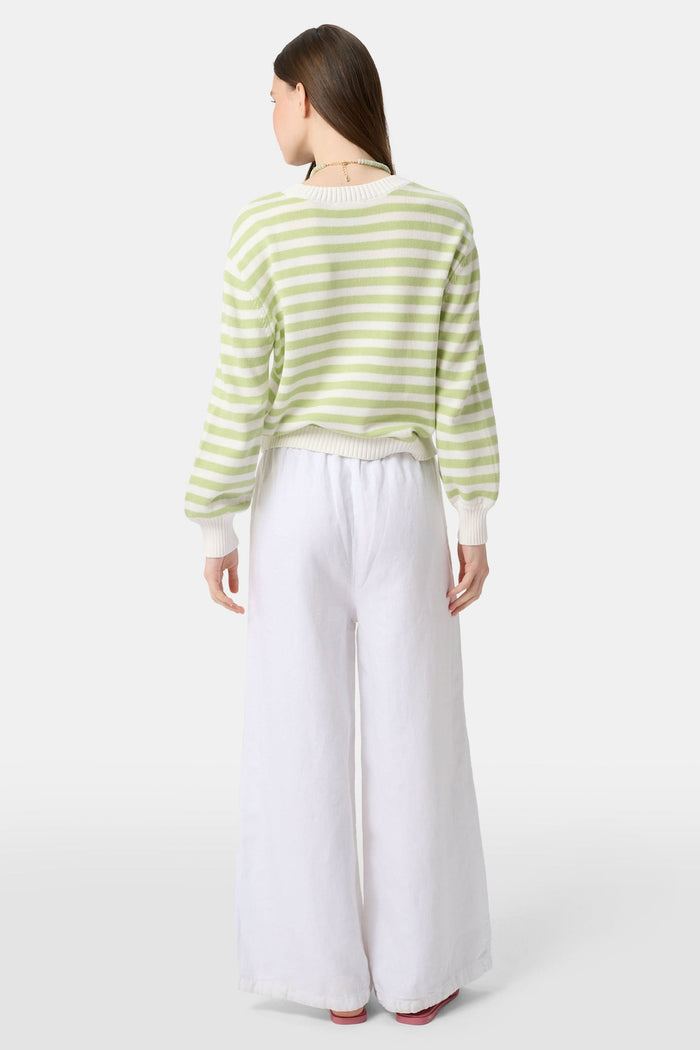 A woman with straight brown hair wears the Roller Rabbit Striped Hadley Sweater in sage green and white stripes, paired with white high-waisted pants and a beaded necklace, standing against a plain background and looking at the camera.