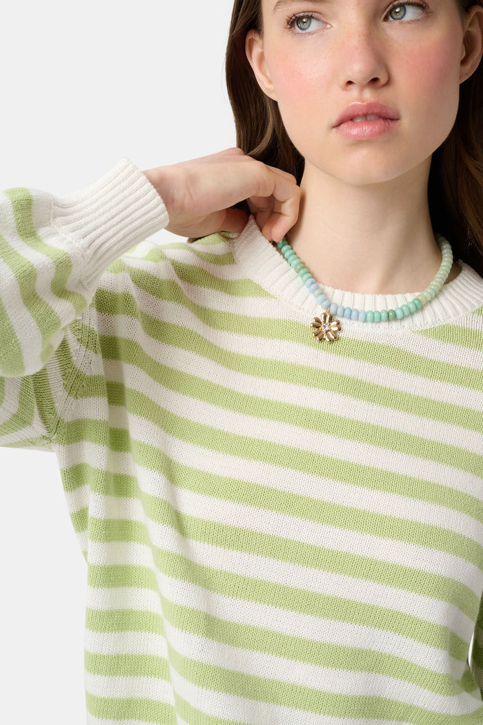 A woman with straight brown hair wears the Roller Rabbit Striped Hadley Sweater in sage green and white stripes, paired with white high-waisted pants and a beaded necklace, standing against a plain background and looking at the camera.