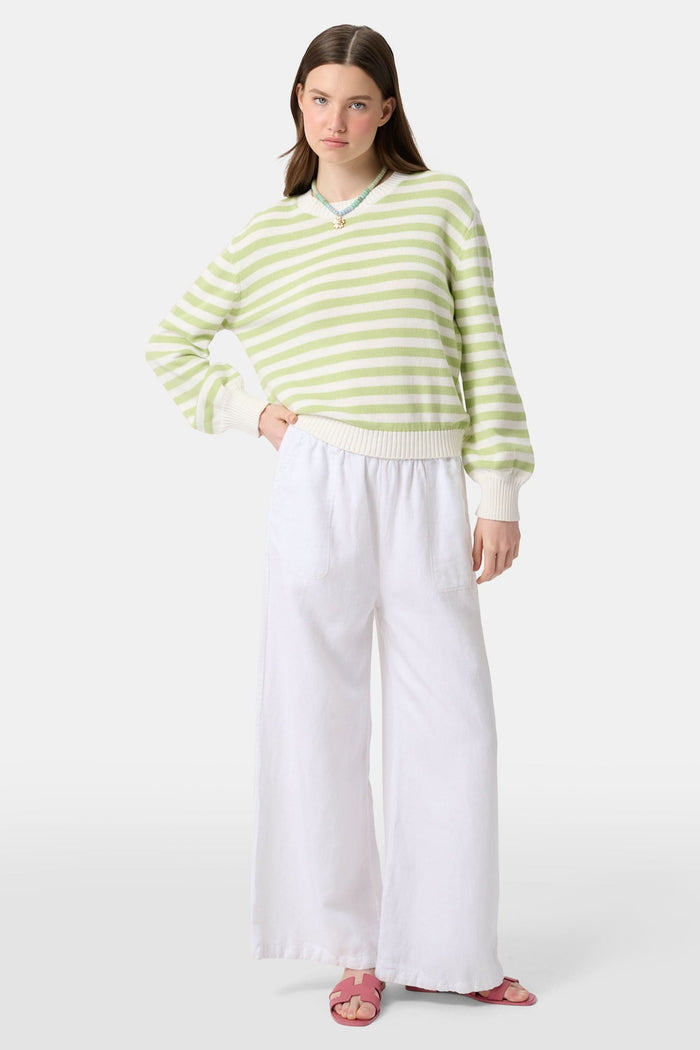 A woman with straight brown hair wears the Roller Rabbit Striped Hadley Sweater in sage green and white stripes, paired with white high-waisted pants and a beaded necklace, standing against a plain background and looking at the camera.