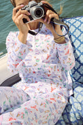 Wearing Roller Rabbit’s Summer Club Pajamas, a person sits on a boat with a blue patterned cushion, holding a camera up for a photo as water glistens in the background.