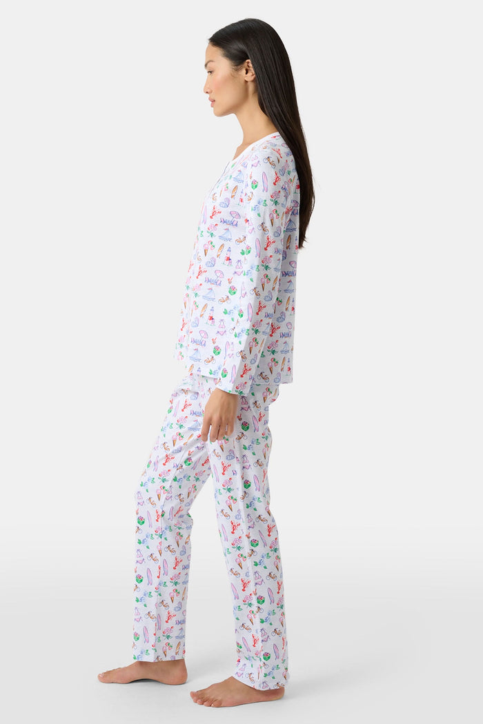 Wearing Roller Rabbit’s Summer Club Pajamas, a person sits on a boat with a blue patterned cushion, holding a camera up for a photo as water glistens in the background.