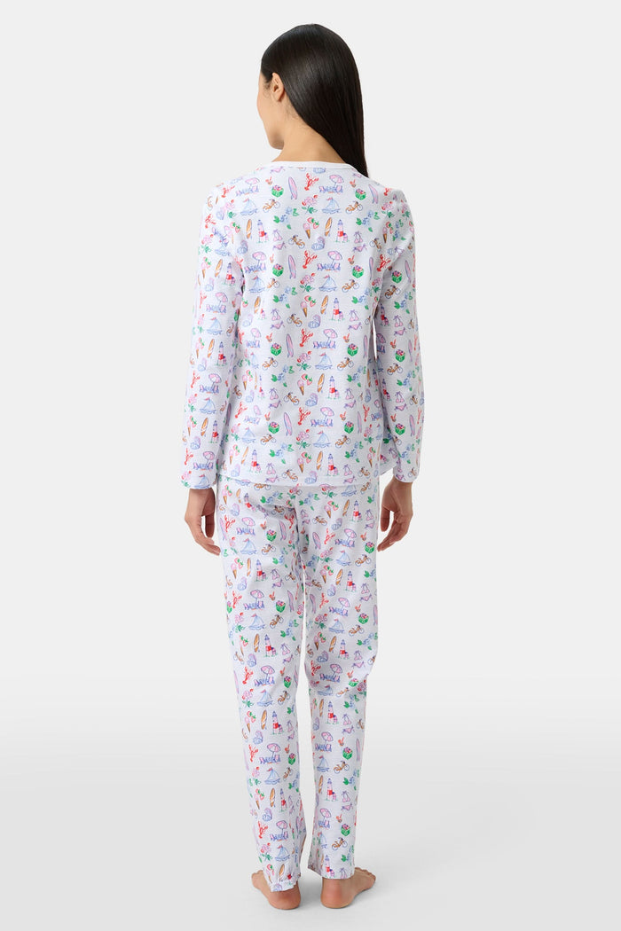 Wearing Roller Rabbit’s Summer Club Pajamas, a person sits on a boat with a blue patterned cushion, holding a camera up for a photo as water glistens in the background.