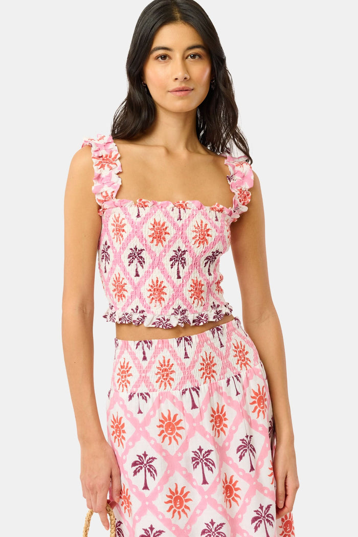 A woman wears the Roller Rabbit Sunny Days Sabrina Top, a cropped, sleeveless ruffled pink and white piece paired with a matching high-waisted skirt in a pink and orange geometric floral linen-viscose blend, standing before a plain light background.