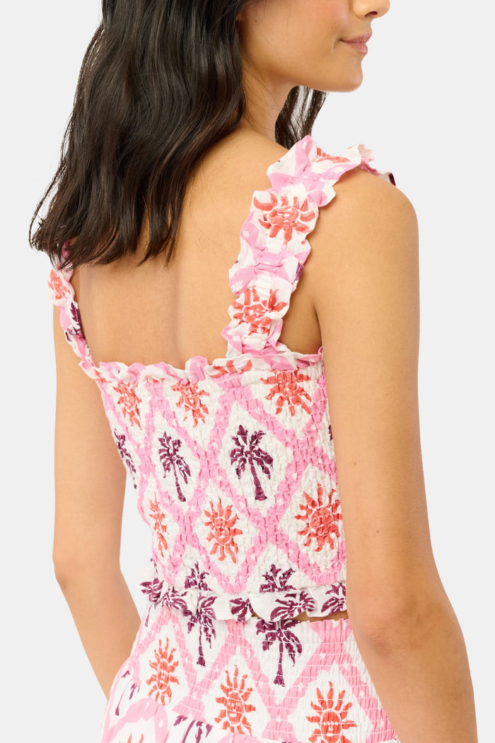 A woman wears the Roller Rabbit Sunny Days Sabrina Top, a cropped, sleeveless ruffled pink and white piece paired with a matching high-waisted skirt in a pink and orange geometric floral linen-viscose blend, standing before a plain light background.