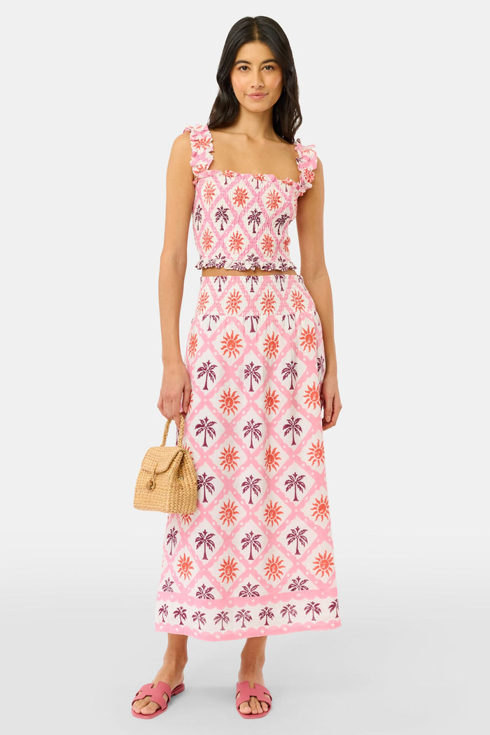 A woman wears the Roller Rabbit Sunny Days Sabrina Top, a cropped, sleeveless ruffled pink and white piece paired with a matching high-waisted skirt in a pink and orange geometric floral linen-viscose blend, standing before a plain light background.