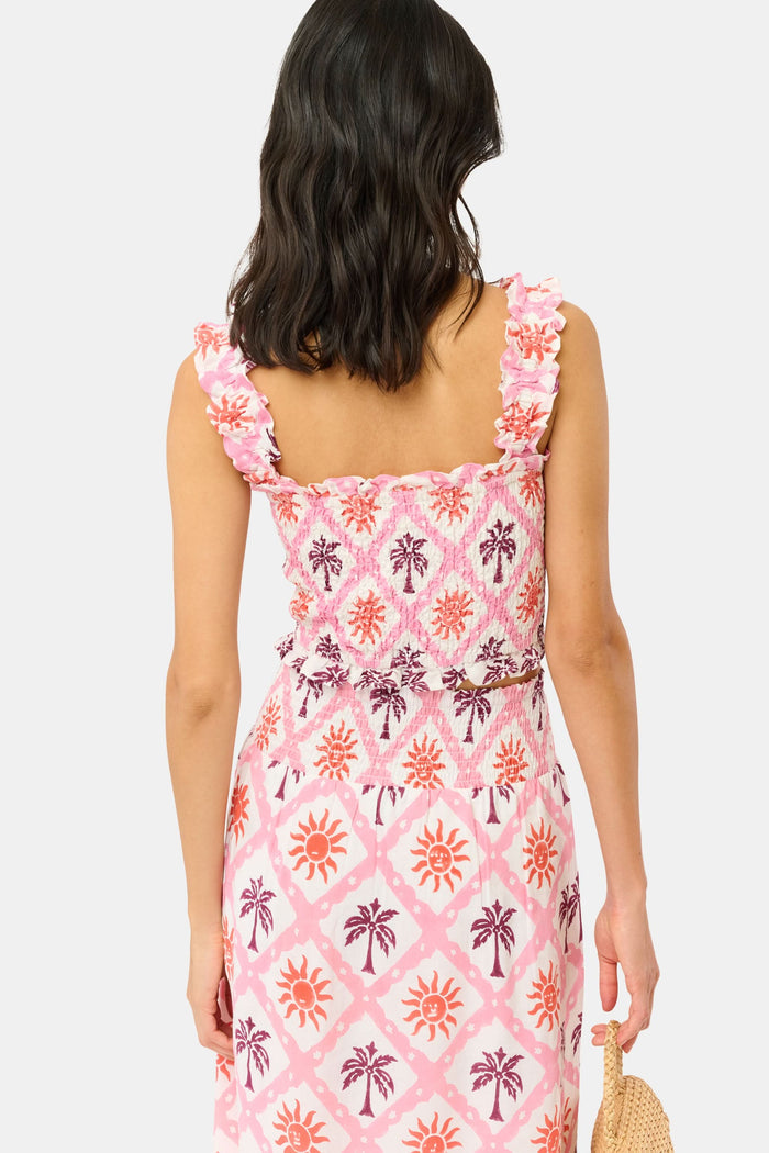 A woman wears the Roller Rabbit Sunny Days Sabrina Top, a cropped, sleeveless ruffled pink and white piece paired with a matching high-waisted skirt in a pink and orange geometric floral linen-viscose blend, standing before a plain light background.