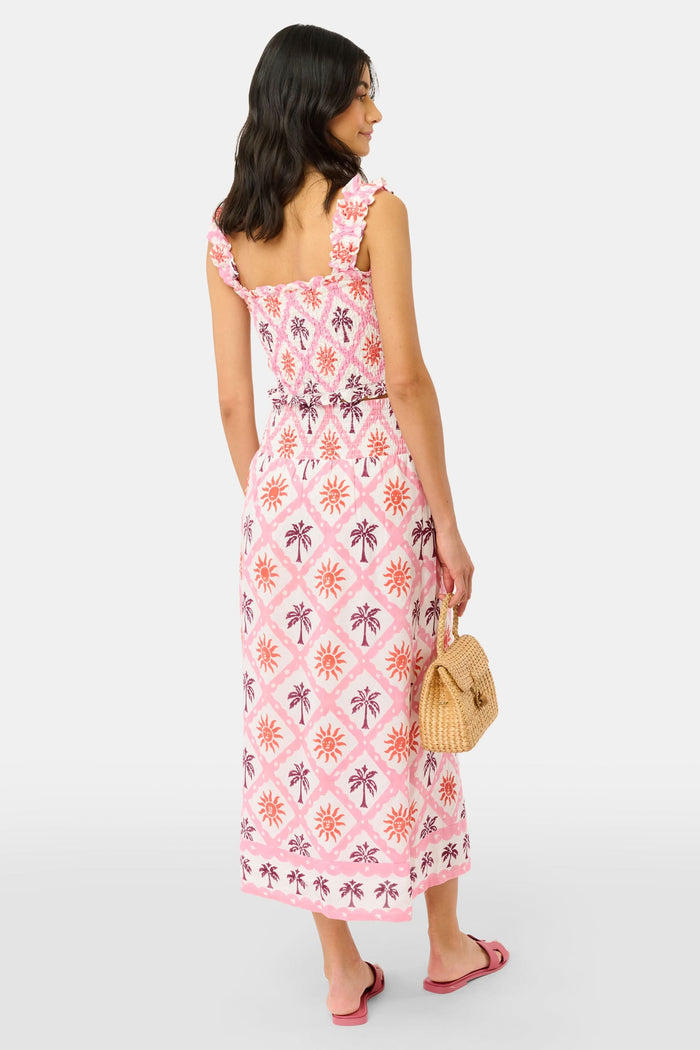 Only the lower half of the body is shown: someone wears the Roller Rabbit Sunny Days Juliette Skirt—a pink and white linen maxi skirt with sun and palm patterns—paired with pink sandals and a small woven handbag.