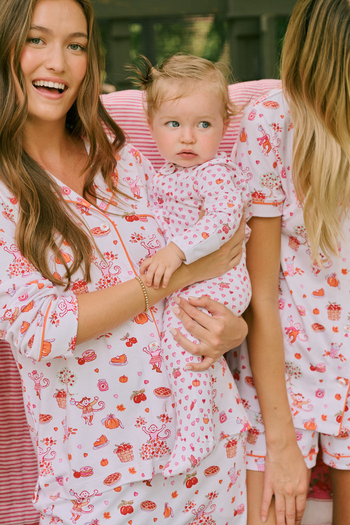A woman stands barefoot against a plain white background, hands in pockets, wearing the Roller Rabbit Sweater Weather Long Sleeve Polo Pajamas—a floral pink and white set featuring a long-sleeved polo top and matching pants.