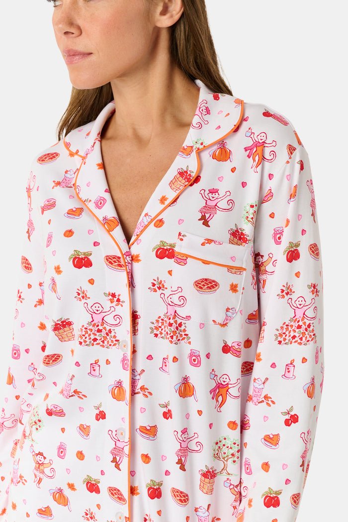 A woman stands barefoot against a plain white background, hands in pockets, wearing the Roller Rabbit Sweater Weather Long Sleeve Polo Pajamas—a floral pink and white set featuring a long-sleeved polo top and matching pants.