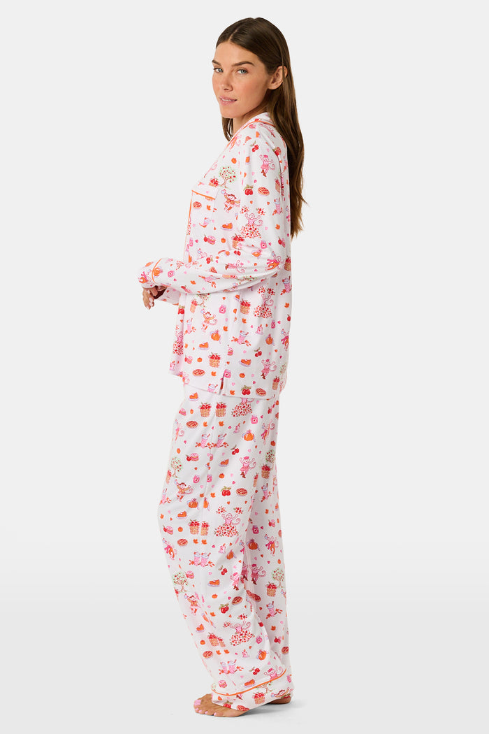A woman stands barefoot against a plain white background, hands in pockets, wearing the Roller Rabbit Sweater Weather Long Sleeve Polo Pajamas—a floral pink and white set featuring a long-sleeved polo top and matching pants.