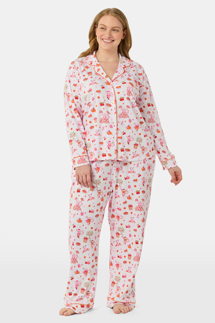 A woman stands barefoot against a plain white background, hands in pockets, wearing the Roller Rabbit Sweater Weather Long Sleeve Polo Pajamas—a floral pink and white set featuring a long-sleeved polo top and matching pants.