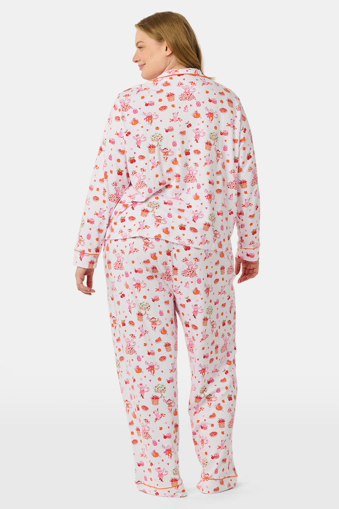 A woman stands barefoot against a plain white background, hands in pockets, wearing the Roller Rabbit Sweater Weather Long Sleeve Polo Pajamas—a floral pink and white set featuring a long-sleeved polo top and matching pants.