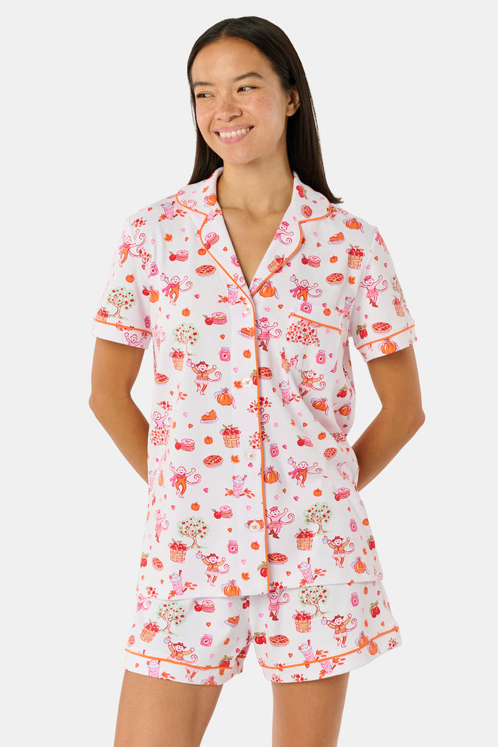 A woman smiles while wearing the Roller Rabbit Sweater Weather Polo Pajamas, a matching set of printed Pima PJs featuring colorful pink and orange cupcakes, flowers, and playful designs on a white background.