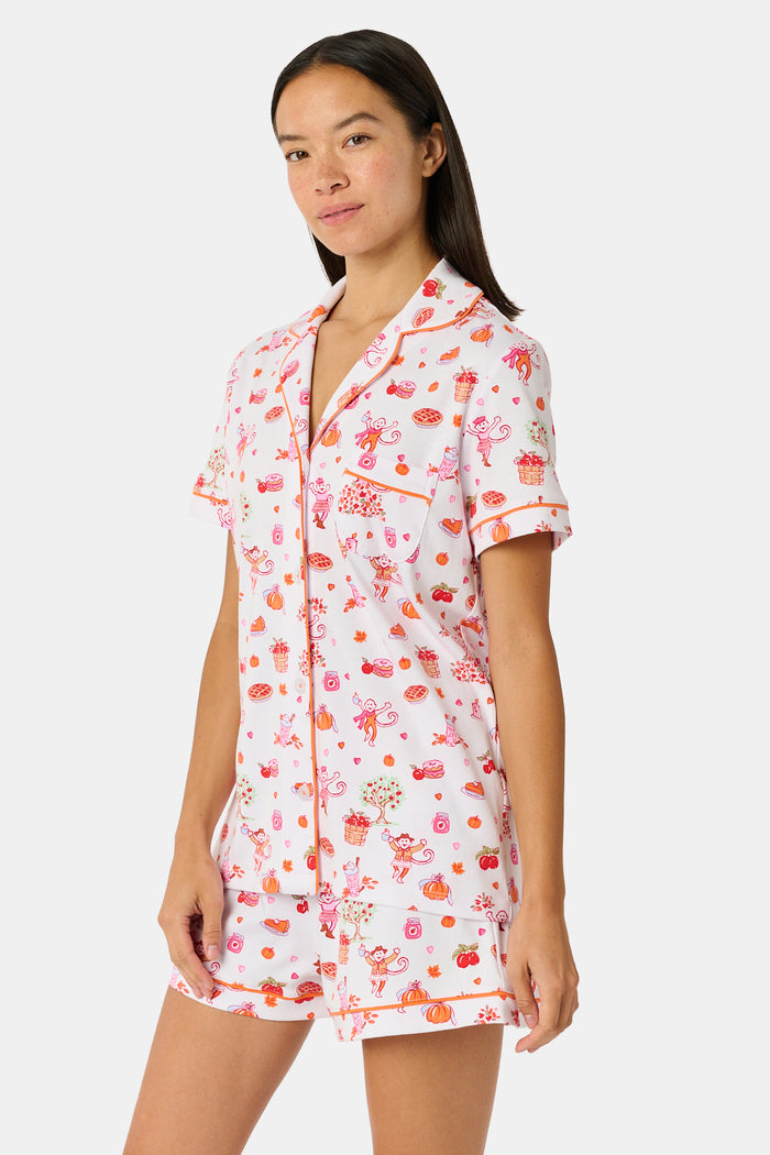 A woman smiles while wearing the Roller Rabbit Sweater Weather Polo Pajamas, a matching set of printed Pima PJs featuring colorful pink and orange cupcakes, flowers, and playful designs on a white background.