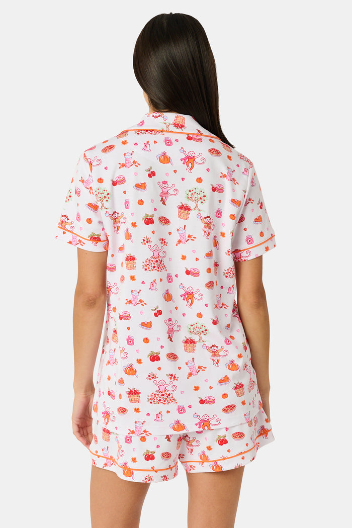 A woman smiles while wearing the Roller Rabbit Sweater Weather Polo Pajamas, a matching set of printed Pima PJs featuring colorful pink and orange cupcakes, flowers, and playful designs on a white background.