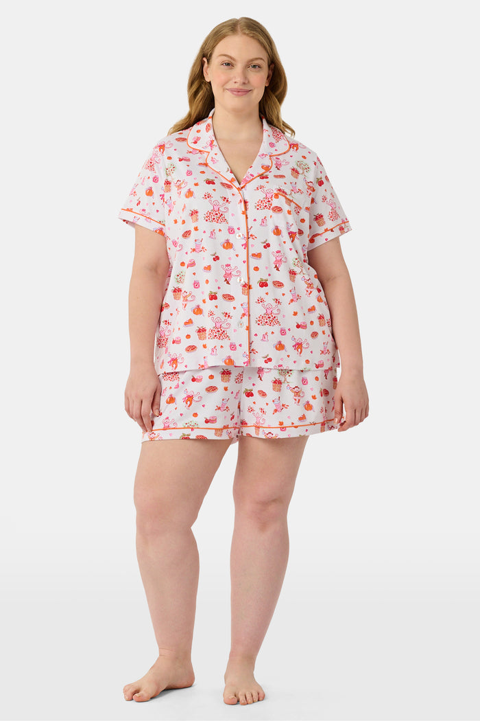 A woman smiles while wearing the Roller Rabbit Sweater Weather Polo Pajamas, a matching set of printed Pima PJs featuring colorful pink and orange cupcakes, flowers, and playful designs on a white background.