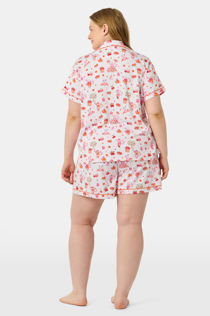 A woman smiles while wearing the Roller Rabbit Sweater Weather Polo Pajamas, a matching set of printed Pima PJs featuring colorful pink and orange cupcakes, flowers, and playful designs on a white background.