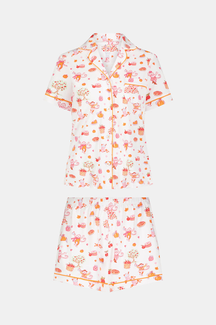 A woman smiles while wearing the Roller Rabbit Sweater Weather Polo Pajamas, a matching set of printed Pima PJs featuring colorful pink and orange cupcakes, flowers, and playful designs on a white background.