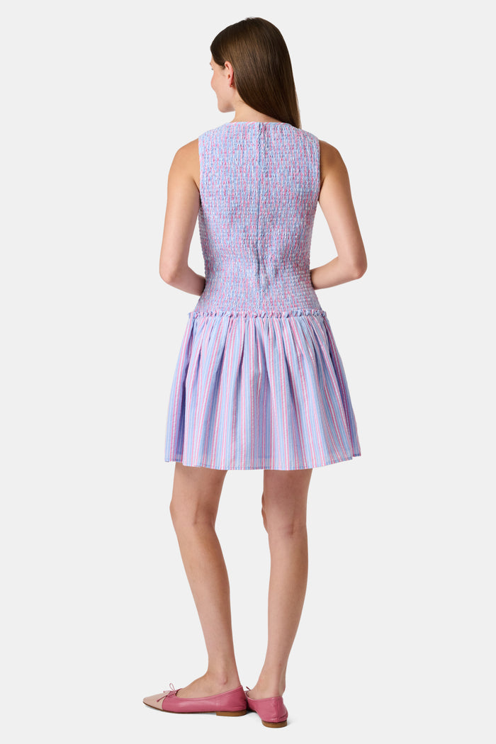 A young girl wearing the Roller Rabbit Kids Sweet Stripe Casey Dress stands in front of lockers, smiling and holding a folder decorated with colorful raindrops.