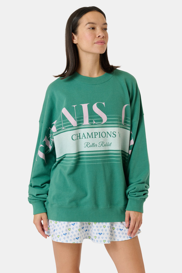 A person wearing a preppy green Roller Rabbit Tennis Champions Andie Sweatshirt stands in front of bookshelves. Only the lower part of their face is visible, and their hands are in their pockets.