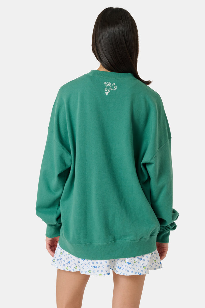 A person wearing a preppy green Roller Rabbit Tennis Champions Andie Sweatshirt stands in front of bookshelves. Only the lower part of their face is visible, and their hands are in their pockets.