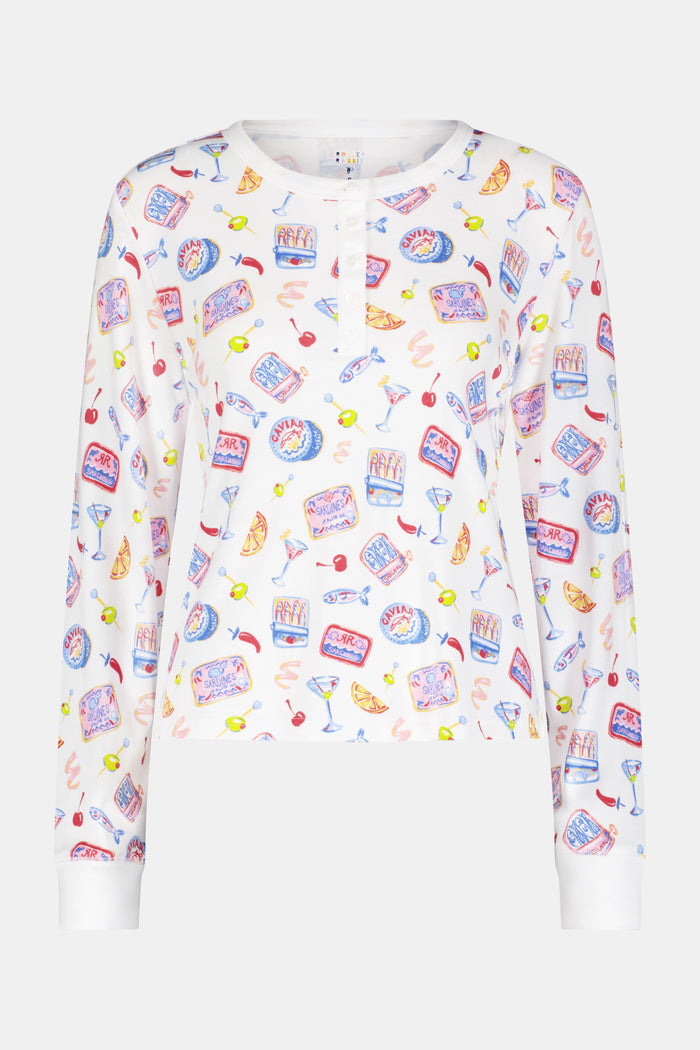 A person wears the Roller Rabbit Tinned or Twist Ashley Top, a white long-sleeve with colorful food and drink prints, while sitting on a couch with red and green patterned pillows in the background. The image is cropped at the shoulders.