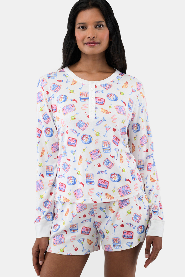 A person wears the Roller Rabbit Tinned or Twist Ashley Top, a white long-sleeve with colorful food and drink prints, while sitting on a couch with red and green patterned pillows in the background. The image is cropped at the shoulders.